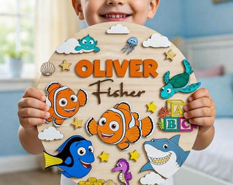 May include: A circular wooden sign featuring the name Oliver Fisher in orange and brown text. The design includes colorful cutouts of clownfish, a blue tang, a shark, a seahorse, a sea turtle, seashells, yellow stars, white clouds, and alphabet blocks.