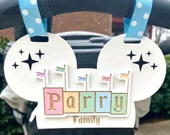 May include: A white, mouse-ear shaped stroller sign featuring the text "Parry Family" in gold lettering. The design includes pastel-colored blocks with small flags and black star icons. It is attached to a stroller handle with blue polka-dot ribbons.