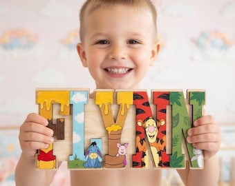 Może przedstawiać: Drewniana układanka z imieniem "FLYNN" w kolorowych literach. Litery ozdobione są postaciami z Kubusia Puchatka i detalami spływającego miodu. Układanka trzymana jest przez uśmiechnięte dziecko.