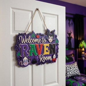 以下が含まれることがあります： 「Welcome to Raven Room」とカラフルな文字で書かれた紫と白の木製サイン。マレフィセントやアースラなど、ディズニーの悪役のイラストが描かれており、白いドアに掛けられています。