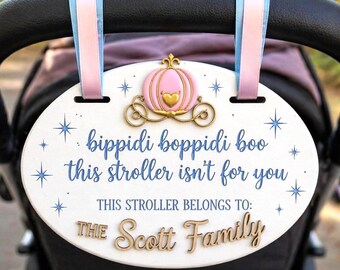 May include: An oval white stroller sign with pink and blue ribbons, featuring a gold pumpkin carriage icon, blue star accents, and the text: bippidi boppidi boo this stroller isn't for you, THIS STROLLER BELONGS TO: THE Scott Family.