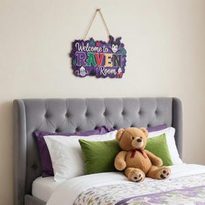 以下が含まれることがあります： 紫、緑、黄色の「Welcome to Raven Room」の文字が入った装飾用の木製サイン。灰色のヘッドボード、紫と緑の枕、テディベアのあるベッドの上にサインが吊り下げられています。