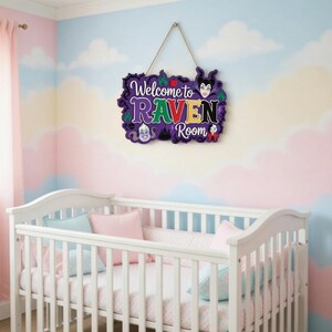 以下が含まれることがあります： 白いベビーベッドの上に吊るされた、さまざまな色の「Welcome to RAVEN Room」というテキストが入った紫色の木製サイン。サインには漫画のキャラクターのイラストが描かれており、天然繊維のロープで吊るされています。保育園の壁はパステルカラーの雲のデザインです。