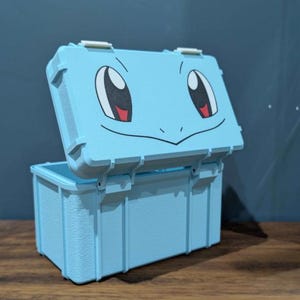 May include: A light blue rectangular storage box with a hinged lid. The lid features a printed graphic of a cartoon face with large white eyes, black pupils, and a curved mouth line. The box has a textured surface and vertical structural ribs.