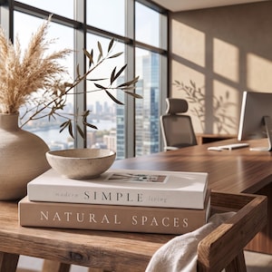 May include: A beige ceramic vase with dried pampas grass and olive branches, a small speckled bowl, and two stacked books titled modern SIMPLE living and NATURAL SPACES, all resting on a wooden surface in a bright office setting.