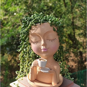 May include: A light peach-colored planter shaped like a human head with closed eyes and pink cheeks. The figure holds a small white cup and saucer. A trailing string of pearls plant cascades over the head, mimicking hair, resting on a round base.