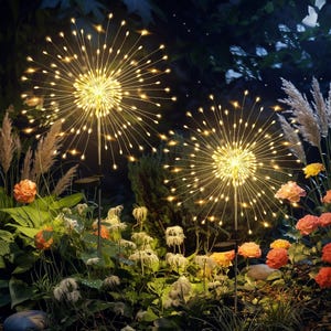 May include: Two solar-powered garden lights featuring a spherical firework design with thin, radiating wire branches tipped with small, warm white LED lights, mounted on slender metal stakes positioned among various green plants and flowers.