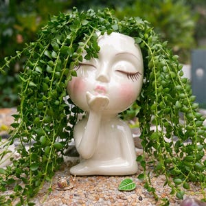 May include: A glossy white ceramic planter shaped like a human head with closed eyes and pursed lips, featuring a hand raised to the mouth in a blowing kiss gesture. The planter is filled with a trailing green succulent plant with small leaves.