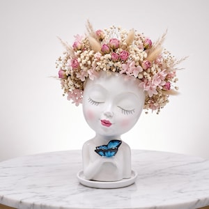 May include: A white ceramic head-shaped planter featuring a serene face with closed eyes and pink cheeks. The top is adorned with a crown of dried pink roses, white baby's breath, and pampas grass. A small blue butterfly rests on the figure's hand.