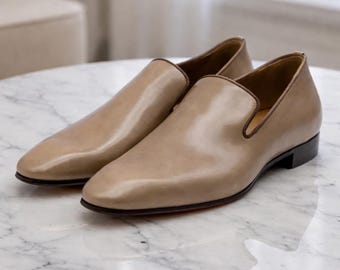May include: A pair of tan leather loafers with a classic design. The shoes have a rounded toe, a low heel, and a dark brown trim. The soles are a dark color. The shoes are displayed on a marble surface.