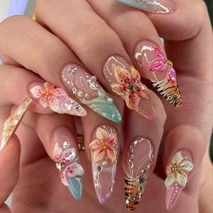 May include: A set of long, almond-shaped acrylic nails. Each nail features a unique design, including 3D floral accents, rhinestones, and tiger stripe patterns. Colors include pink, peach, blue, and clear.