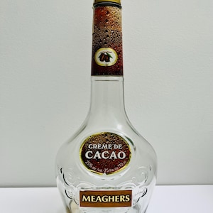 May include: A clear glass bottle of Meagher's Creme de Cacao liqueur. The bottle has a gold cap and a brown label with the text "Creme de Cacao" and "25% alc./vol. 25 fl.oz./710 ml".