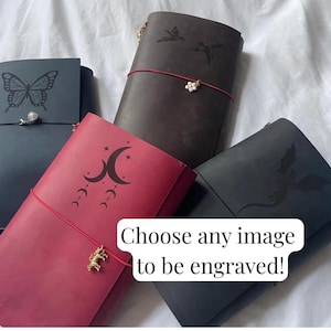 Op de afbeelding: Vier leren notitieboeken in zwart, donkerbruin en rood, elk met unieke lasergegraveerde ontwerpen zoals een vlinder, vogels, een maanfase en een draak. Elk notitieboek is vastgezet met een elastische band en een bedeltje. Tekst: Choose any image to be engraved!