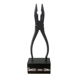 Könnte beinhalten: Eine schwarze Metallzangen-Skulptur auf einem schwarzen Holzsockel. Die Zange ist geöffnet, und der Sockel trägt den weißen Text "Pince - sans - Rire". Die Skulptur ist ein dekoratives Objekt.