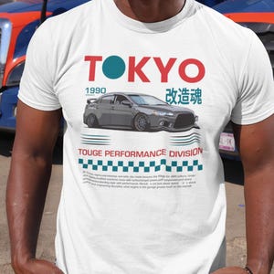 May include: White t-shirt with a graphic design featuring the word "TOKYO" in red and a teal circle, a gray car illustration, and Japanese characters. The shirt also includes the text "TOUGE PERFORMANCE DIVISION".