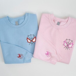 May include: Two folded sweatshirts, one light blue and one pink. Each has a Spider-Man mask embroidered on the chest and a spider on the sleeve. The light blue mask is red and black, and the pink mask is pink and black.