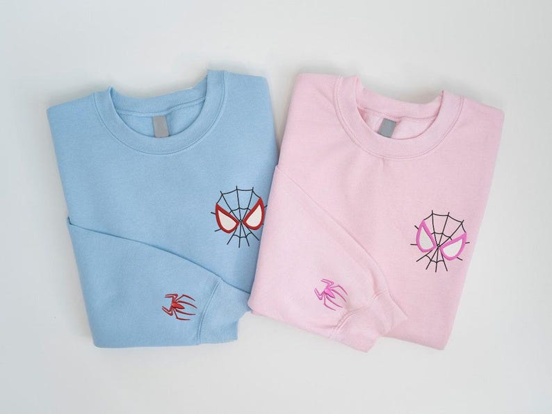 May include: Two folded sweatshirts, one light blue and one pink. Each has a Spider-Man mask embroidered on the chest and a spider on the sleeve. The light blue mask is red and black, and the pink mask is pink and black.