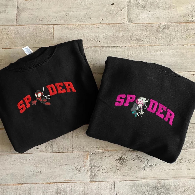 Puede incluir: Dos sudaderas negras. Una presenta la palabra "SPIDER" en rojo, con un gr&aacute;fico de ara&ntilde;a roja y negra. La otra tiene la palabra "SPIDER" en rosa, con un gr&aacute;fico de ara&ntilde;a blanca, negra y turquesa.