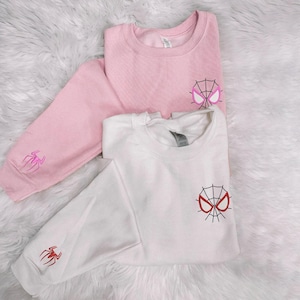 May include: Two sweatshirts, one pink and one white, are displayed on a fluffy white surface. Each sweatshirt features a spider web design on the chest and a spider emblem on the sleeve. The pink sweatshirt has a pink spider web design, while the white one has a red spider web design.