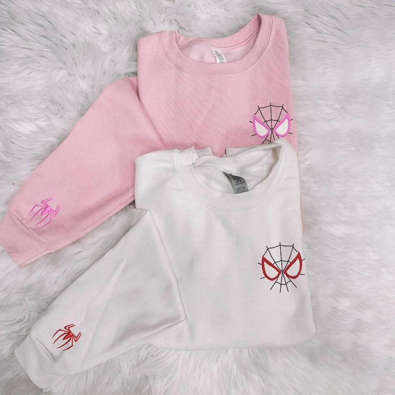 May include: Two sweatshirts, one pink and one white, are displayed on a fluffy white surface. Each sweatshirt features a spider web design on the chest and a spider emblem on the sleeve. The pink sweatshirt has a pink spider web design, while the white one has a red spider web design.