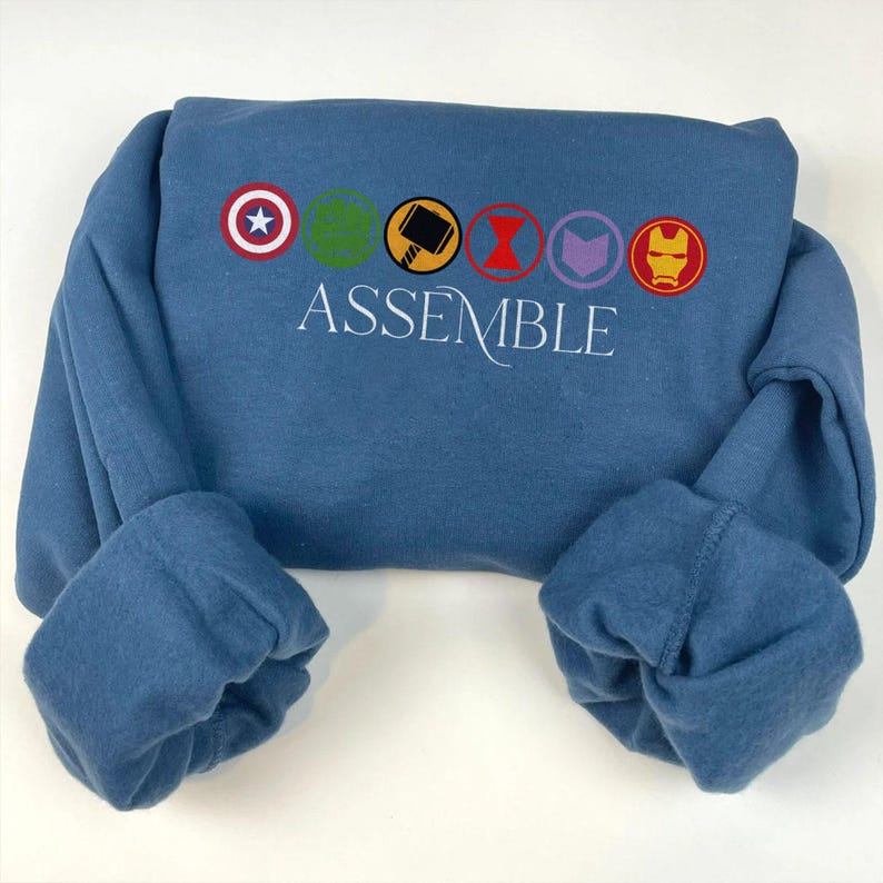 May include: A blue sweatshirt featuring the word "ASSEMBLE" in white script. Above the word are colourful circular emblems of Captain America, Hulk, Thor, Black Widow, Hawkeye, and Iron Man.