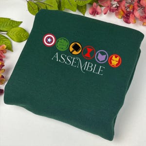 May include: Dark green sweatshirt featuring the word "ASSEMBLE" in white script. Above the word are six embroidered circular emblems. The emblems include a star, a fist, a hammer, a red hourglass, a purple arrow, and a gold mask.