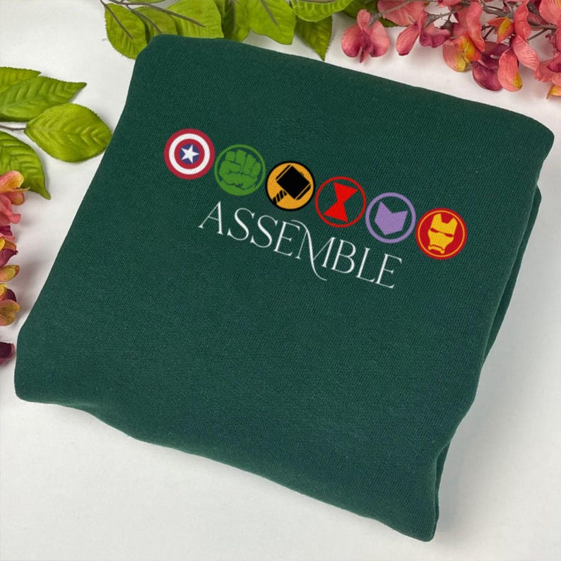 May include: Dark green sweatshirt featuring the word "ASSEMBLE" in white script. Above the word are six embroidered circular emblems. The emblems include a star, a fist, a hammer, a red hourglass, a purple arrow, and a gold mask.