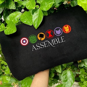 May include: Black sweatshirt with embroidered superhero symbols and the word "ASSEMBLE". The symbols are in a circle and include a star, a fist, a hammer, an hourglass, an arrow, and a mask. The sweatshirt is held against green leaves.