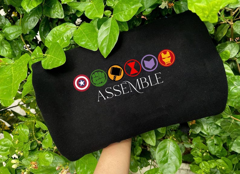May include: Black sweatshirt with embroidered superhero symbols and the word "ASSEMBLE". The symbols are in a circle and include a star, a fist, a hammer, an hourglass, an arrow, and a mask. The sweatshirt is held against green leaves.