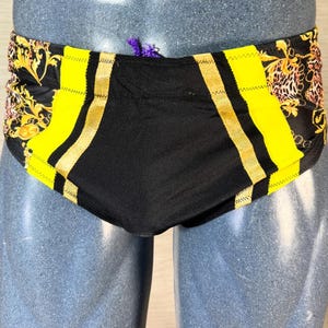 May include: Black and yellow swim trunks with gold accents. The trunks feature a black center panel with yellow side panels and gold stripes. The waistband has a decorative pattern with gold and black floral and animal print.