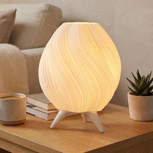 Puede incluir: Una lámpara de mesa blanca en forma de huevo con un diseño texturizado en espiral, apoyada sobre tres patas blancas. La lámpara está encendida, emitiendo un brillo cálido. Una pequeña suculenta en maceta y una taza de café están cerca.