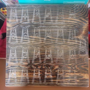 May include: A square wooden board with a dark, stained wood grain finish featuring twenty-three laser-engraved outlines of capes. Each cape outline contains a unique icon representing various skills, arranged in a grid pattern across the surface.