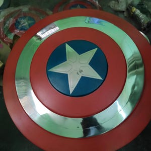 Op de afbeelding: Een rood, wit en blauw Captain America schild. Het schild heeft een rode buitenring, een zilveren binnenring en een blauw midden met een witte ster. Andere schilden zijn zichtbaar op de achtergrond.
