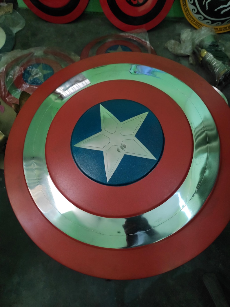 Op de afbeelding: Een rood, wit en blauw Captain America schild. Het schild heeft een rode buitenring, een zilveren binnenring en een blauw midden met een witte ster. Andere schilden zijn zichtbaar op de achtergrond.