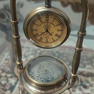 Puede incluir: Reloj de bolsillo antiguo de latón con soporte. La esfera del reloj muestra "VICTORIA LONDON 1876" con números romanos. El soporte tiene un diseño arqueado con una brújula en la base, de estilo vintage.