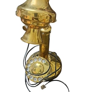 May include: A polished brass candlestick telephone featuring a circular rotary dial with white numbered buttons from zero to nine. The device includes a prominent top-mounted receiver cradle and a thin black coiled cord extending from the base.