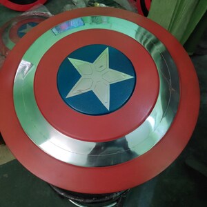 Op de afbeelding: Een Captain America-schild in rood, zilver en blauw. Het schild heeft een rode buitenring, een zilveren binnenring en een blauw midden met een witte ster. Er zijn meerdere schilden zichtbaar.