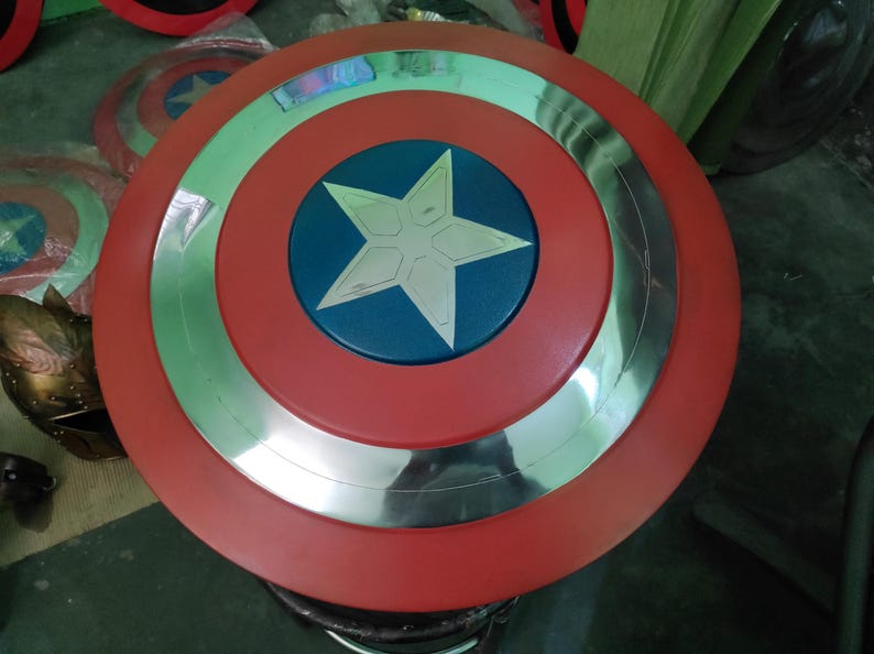 Op de afbeelding: Een Captain America-schild in rood, zilver en blauw. Het schild heeft een rode buitenring, een zilveren binnenring en een blauw midden met een witte ster. Er zijn meerdere schilden zichtbaar.