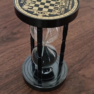 May include: A black hourglass with a circular top featuring a chessboard pattern, chess piece icons, and the text "THE CHESS MAKER 6th CENTURY AD" and "SAND TIMER". The device has four black pillars connecting the top and base, with black sand inside.