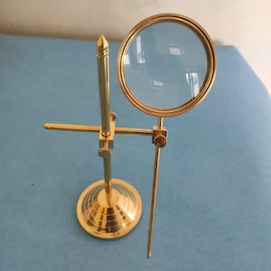 Può includere: Lente d'ingrandimento in ottone con lente rotonda e supporto decorativo. Il supporto ha una base circolare e un braccio regolabile per sostenere la lente d'ingrandimento. La lente d'ingrandimento è uno strumento classico.