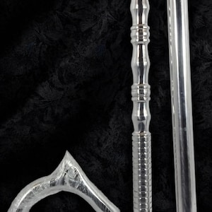 Op de afbeelding: Een zilverkleurige wandelstok gedemonteerd in drie delen: een gebogen handvat verpakt in beschermend plastic, een gestructureerde metalen schacht met decoratieve groeven en een gladde schachtverlenging met een zwarte rubberen dop aan de basis.