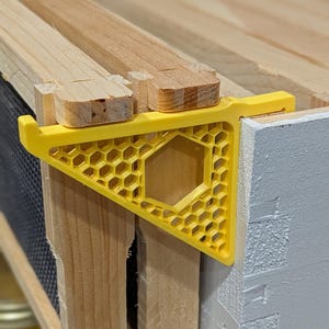 Può includere: Primo piano di un distanziatore per telai di alveare in plastica gialla con un design a nido d'ape. Il distanziatore è posizionato tra i telai dell'alveare in legno. I telai dell'alveare sono in legno chiaro e parzialmente assemblati.