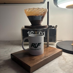 Könnte beinhalten: Ein Kaffeezubereitungssystem mit einem Glasfilter, einer weißen Tasse mit dem Text "RUF HIGHSNOBIETY" und einem Holzsockel. Kaffee tropft in die Tasse. Eine Retro-Uhr befindet sich im Hintergrund.