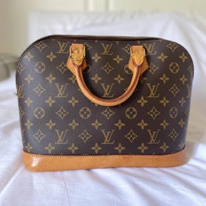 K&ouml;nnte beinhalten: Braune Handtasche mit sich wiederholendem Monogrammmuster. Die Tasche hat eine abgerundete Form, einen hellbraunen Lederboden und zwei Griffe. Goldfarbene Hardware akzentuiert die Griffe und den Rei&szlig;verschluss.