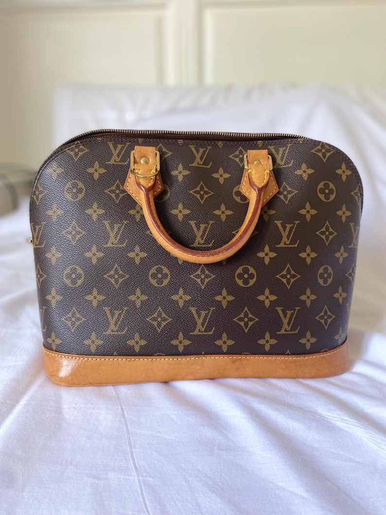 K&ouml;nnte beinhalten: Braune Handtasche mit sich wiederholendem Monogrammmuster. Die Tasche hat eine abgerundete Form, einen hellbraunen Lederboden und zwei Griffe. Goldfarbene Hardware akzentuiert die Griffe und den Rei&szlig;verschluss.
