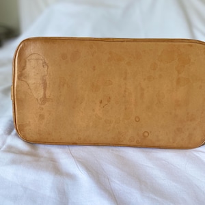 K&ouml;nnte beinhalten: Nahaufnahme des Bodens einer hellbraunen Lederhandtasche. Das Leder weist dunkelbraune Wasserflecken und -punkte auf. Die Tasche hat abgerundete Ecken und eine glatte Oberfl&auml;che.