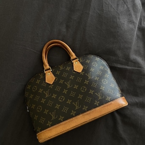 K&ouml;nnte beinhalten: Braune Handtasche mit abgerundeter Form und einem sich wiederholenden Muster aus ineinandergreifenden Logos. Die Tasche hat braune Lederbes&auml;tze und -griffe sowie goldfarbene Hardware-Akzente. Der Hintergrund ist dunkel.