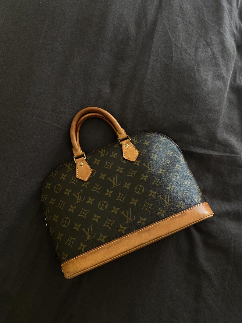 K&ouml;nnte beinhalten: Braune Handtasche mit abgerundeter Form und einem sich wiederholenden Muster aus ineinandergreifenden Logos. Die Tasche hat braune Lederbes&auml;tze und -griffe sowie goldfarbene Hardware-Akzente. Der Hintergrund ist dunkel.