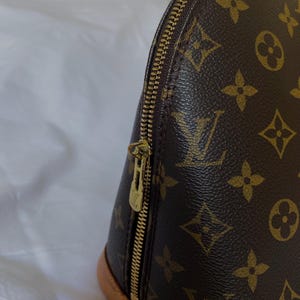 K&ouml;nnte beinhalten: Nahaufnahme einer braunen Handtasche mit goldenem Rei&szlig;verschluss. Die Tasche hat ein sich wiederholendes Muster aus braunen Blumen- und Logodesigns. Der Boden der Tasche ist hellbraun.