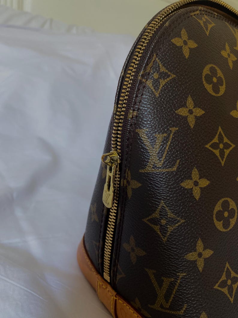 K&ouml;nnte beinhalten: Nahaufnahme einer braunen Handtasche mit goldenem Rei&szlig;verschluss. Die Tasche hat ein sich wiederholendes Muster aus braunen Blumen- und Logodesigns. Der Boden der Tasche ist hellbraun.